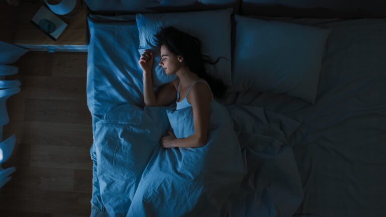 Top View of Beautiful Young Woman Sleeping Cozily on a Bed in His Bedroom at Night. Blue Nightly Colors with Cold Weak Lamppost Light Shining Through the Window