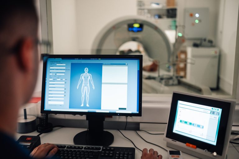 A technician is observed monitoring patient data on a computer during the CT scan process, while the CT scanner is in operation in the background. The control room is equipped with screens and medical devices.