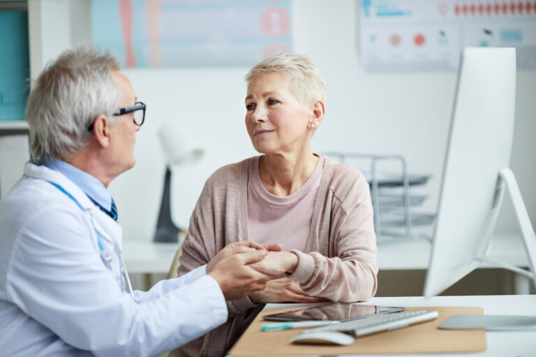 Senior doctor in eyeglasses sitting at table and holding hand of mature female patient while giving hope and comfort to her.