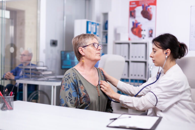 Focused young cardiologist listening heartbeat of senior woman patient at checkup meeting in clinic office.